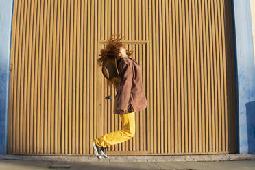 Junge Frau mit zerzaustem Haar, die in der Nähe einer orangefarbenen Metallwand springt - MGRF01241