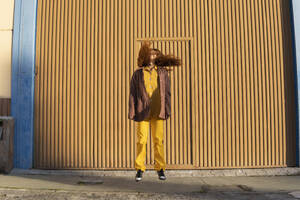 Young woman with tousled hair jumping in front of orange metal wall - MGRF01242