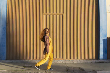 Lächelnde junge Frau mit Jacke auf dem Fußweg in der Nähe einer orangefarbenen Metallwand - MGRF01245