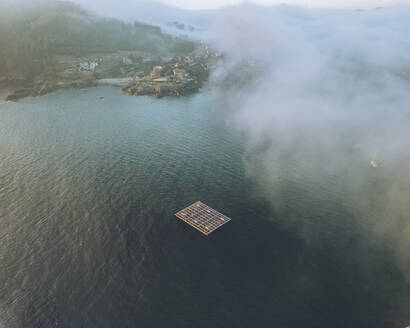 Aerial view of Galicia's coastal town Monteferro with misty morning fog, mussel trawlers and marine farming, Nigran, Rias Baixas, Spain. - AAEF28506