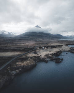 Aerial view of a solitary road winding toward a coastal mountain under overcast skies, Austurland, Iceland. - AAEF28777