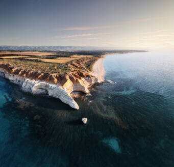 Aerial view of rugged limestone cliffs with a turquoise blue ocean in the foreground and a white sandy beach at sunset, Port Willunga, South Australia, Australia. - AAEF29041