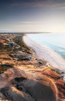 Luftaufnahme einer Klippe mit einer Person in einer orangefarbenen Jacke, die auf dem Gipfel steht, und türkisblauem Meer und weißem Sandstrand bei Sonnenuntergang, Port Willunga, Südaustralien, Australien. - AAEF29047