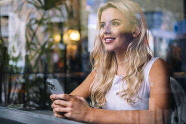 Smiling young woman sitting with smart phone seen through glass - AMWF02088