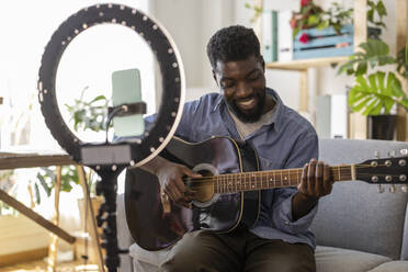 Lächelnder Influencer spielt Gitarre und filmt zu Hause mit seinem Smartphone - JCCMF11575