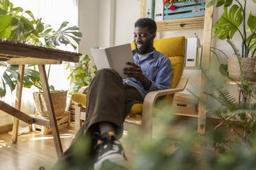 Smiling non-binary person reading book on armchair at home - JCCMF11581