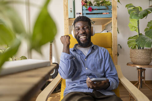 Cheerful non-binary person wearing wireless headphones and sitting on armchair with smart phone at home - JCCMF11583