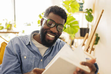 Lächelnde nicht-binäre Person, die eine Brille trägt und ein Buch im heimischen Wohnzimmer liest - JCCMF11608