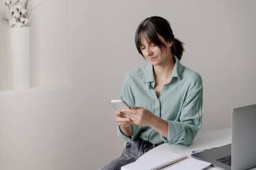 Businesswoman using smart phone sitting near table at home - OGF01310