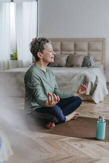 Lächelnde Frau meditiert im Schneidersitz auf einer Yogamatte zu Hause - YTF02093