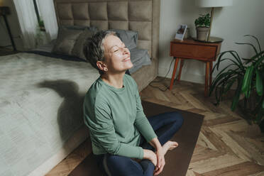 Smiling woman sitting with eyes closed near bed at home - YTF02107