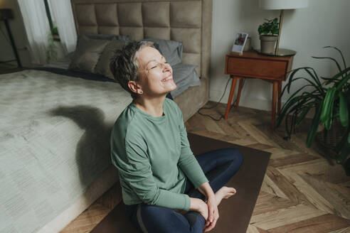 Lächelnde Frau sitzt mit geschlossenen Augen in der Nähe des Bettes zu Hause - YTF02107