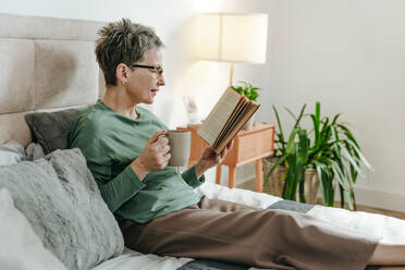 Frau hält Tasse und liest Buch auf dem Bett zu Hause - YTF02114
