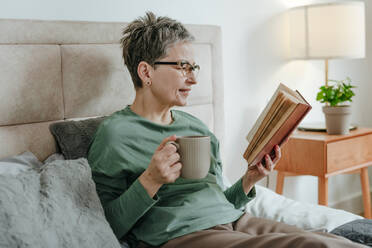 Reife Frau liest Buch auf dem Bett sitzend zu Hause - YTF02115