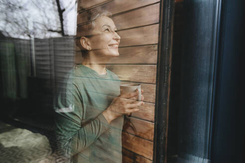 Lächelnde Frau, die eine Kaffeetasse hält und in der Nähe eines Glasfensters steht - YTF02121