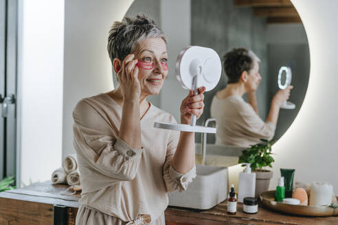 Lächelnde Frau, die eine Augenmaske aufträgt und sich zu Hause im Spiegel betrachtet - YTF02135