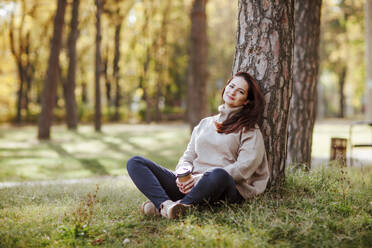 Lächelnde reife Frau hält Kaffeetasse und sitzt im Park - NLAF00387