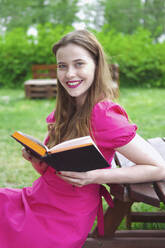 Smiling woman sitting on bench holding book at park - YHF00122