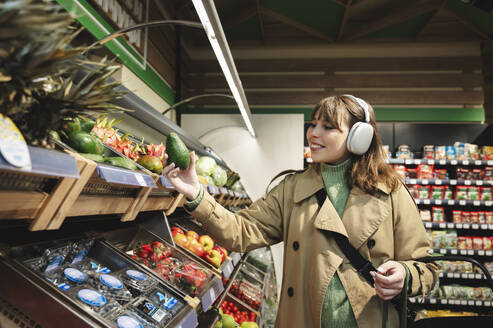 Smiling woman wearing wireless headphones and picking up avocado from rack at store - ALKF01149