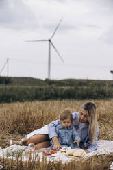 Mother spending leisure time with daughter sitting on blanket at wind farm - KANF00045