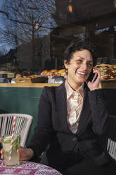 Happy businesswoman holding glass of juice and talking on smart phone at sidewalk cafe - AMWF02095