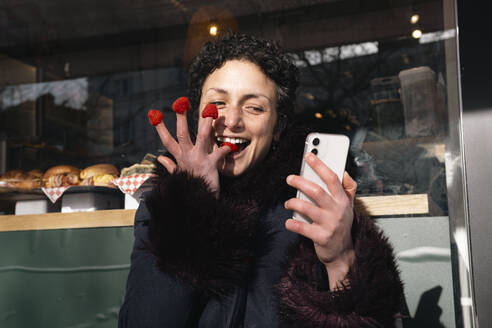 Verspielte Frau isst Himbeeren mit den Fingern und benutzt ein Smartphone in einem Straßencafé - AMWF02100