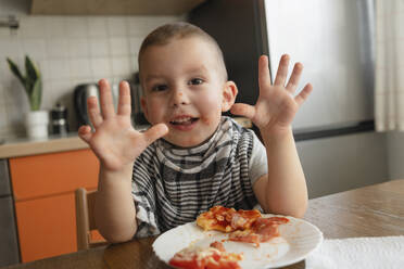 Glücklicher Junge zeigt Hände in der Nähe von Pizza am Tisch in der Küche - KVBF00101