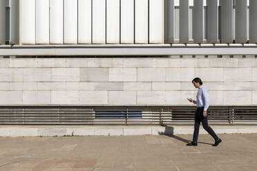 Businessman with smart phone walking on footpath - LMCF01029