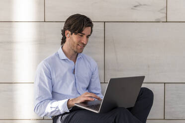 Smiling young businessman using laptop in front of wall - LMCF01036