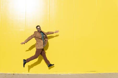 Man posing jumping dressed in an aquamarine shirt and a brown jacket. Yellow background. Granada, Spain. - MGRF01274