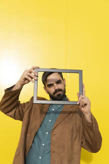 Man wearing an aquamarine shirt and a brown jacket posing with a photo frame. Yellow background. Granada, Spain. - MGRF01280