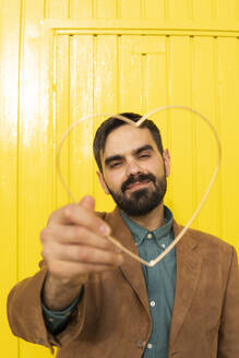 Man wearing an aquamarine shirt and a brown jacket posing with a metal heart. Yellow background. Granada, Spain. - MGRF01289