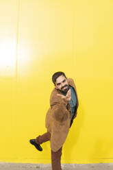 Glücklicher Mann mit brauner Jacke und Gesten vor einer gelben Wand - MGRF01298