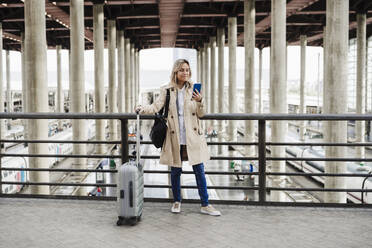 Frau benutzt Smartphone und steht mit Gepäck am Bahnhof - EBBF08834
