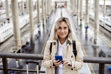 Lächelnde Frau mit gesträhntem Haar, die ein Smartphone am Bahnhof hält - EBBF08840