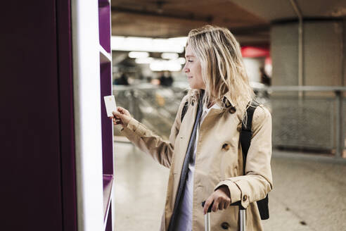 Lächelnde Frau mit Kreditkarte in der Nähe eines Fahrkartenautomaten am Bahnhof - EBBF08859