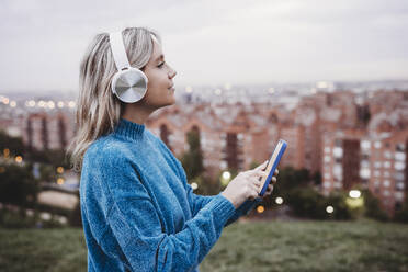 Frau mit kabellosen Kopfhörern hält Smartphone bei Sonnenuntergang - EBBF08895