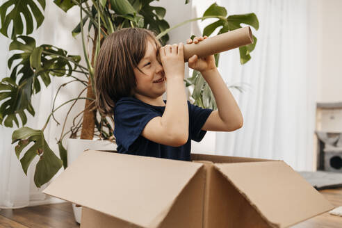 Glücklicher Junge sitzt in einer Schachtel und schaut durch ein Pappteleskop zu Hause - ELMF00170