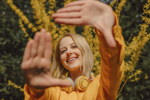 Lächelnde Frau mit gelben drahtlosen Kopfhörern, die durch einen Fingerrahmen schaut - VSNF01824