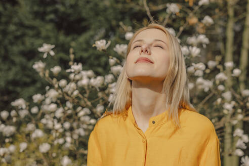 Lächelnde blonde Frau genießt das Sonnenlicht in der Nähe des Magnolienblütenbaums - VSNF01828