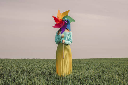 Junge Frau, die ein mehrfarbiges Windrad-Spielzeug hält und in einem grünen Weizenfeld unter dem Himmel steht - VSNF01834