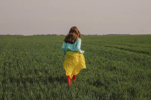 Junge Frau trägt gelben Rock und läuft auf grünem Weizenfeld unter Himmel - VSNF01836