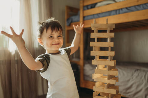 Confident boy with arms raised near wooden block tower at home - ANAF02843