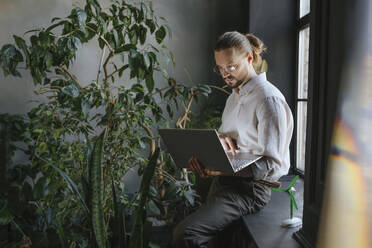 Businessman working on laptop and sitting near window at office - YTF02145