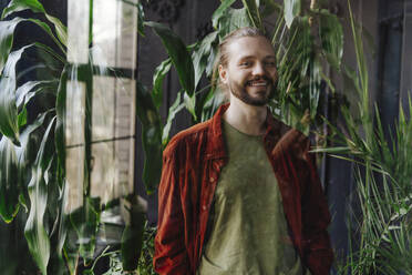 Lächelnder Mann, der in der Nähe von Pflanzen steht, gesehen durch Glas - YTF02178
