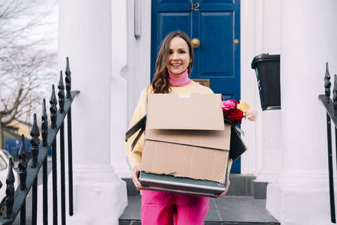 Lächelnde Frau steht mit Kartons vor dem Hauseingang - AMWF02124