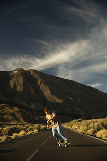 Sporty boy going down a road with a Skate board with the Teide in the background. Tenerife, Canary Islands, Spain. - ACPF01576