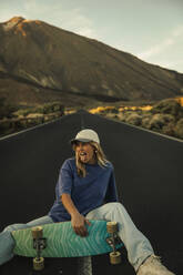 Carefree young woman sitting with skateboard on mountain road - ACPF01578