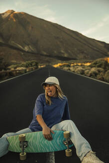 Blonde girl with a cap sitting in the middle of the road posing with a Skateboard with the Teide in the background. Tenerife, Canary Islands, Spain. - ACPF01578