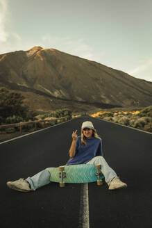 Blonde girl with a cap sitting in the middle of the road posing with a Skateboard with the Teide in the background. Tenerife, Canary Islands, Spain. - ACPF01579